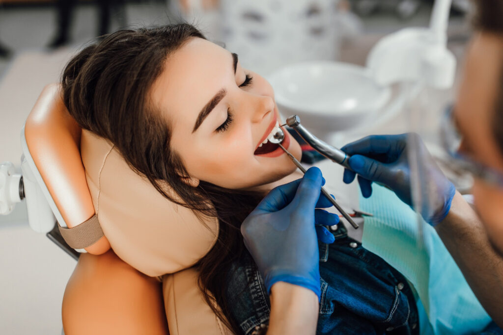 A woman smiling while sitting in a dentist's chair during an oral examination, with a dentist wearing blue gloves using dental instruments to check her teeth