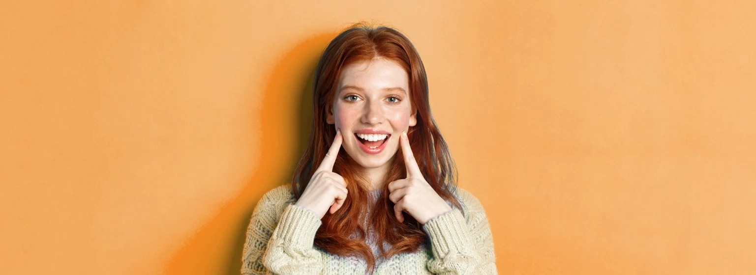 Smiling woman with red hair pointing to her bright white teeth against an orange background.