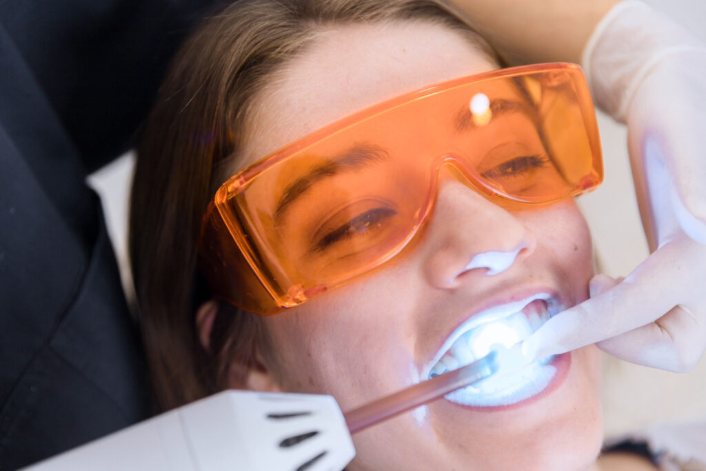 Close-up of a woman wearing orange protective glasses during a teeth whitening session with a blue light device.