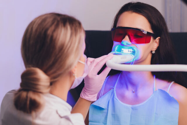 A dentist using a blue light teeth whitening device on a patient wearing protective red glasses and a blue bib.