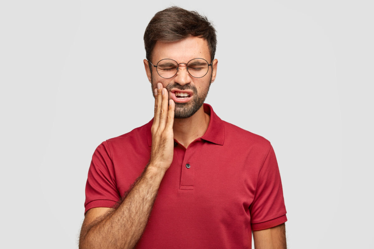 A man wearing glasses and a red polo shirt touching his cheek, expressing pain or discomfort, due to wisdom teeth removal